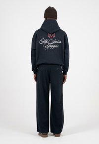 Granatowy hoodie z dużą białą grafiką „Szwajcarski Yuppie” z tyłu. W połączeniu z ciemnymi spodniami o szerokich nogawkach i brązowym obuwiem.
