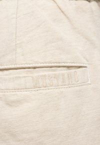 Beżowa bawełniana tkanina o teksturowanej powierzchni. Posiada przeszywaną kieszeń i wytłaczane logo "MUSTANG", co tworzy subtelny detal.