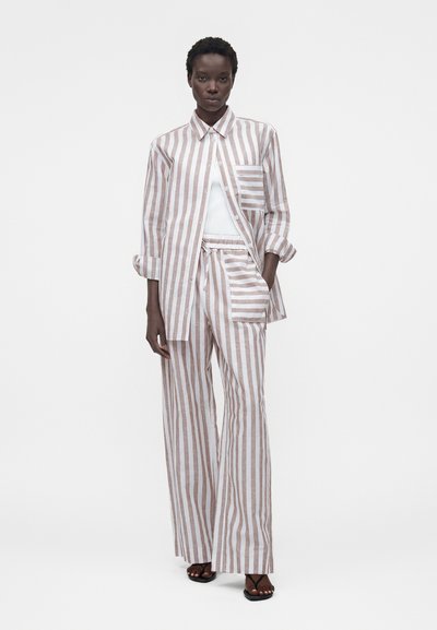 Γυναίκα που φοράει μπεζ και λευκό πουκάμισο με κάθετες ρίγες και ασορτί παντελόνι με φαρδιά μπατζάκια, λευκό τοπ από μέσα, στέκεται με το ένα χέρι στην τσέπη.