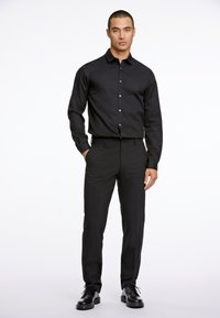 Zwarte langemouwen shirt met knopen, gecombineerd met een aansluitende zwarte broek. Beide zijn gemaakt van een gladde stof. Zwarte veterschoenen maken de look compleet.