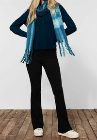 Sciarpa a quadri blu con frange, abbinata a una maglia scura e pantaloni neri a zampa. La modella indossa sneakers bianche, in piedi su un pavimento di legno.