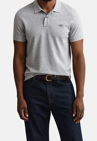 Homme portant un polo gris clair à manches courtes avec un petit logo brodé, un jean bleu foncé et une ceinture tressée marron avec une boucle dorée.