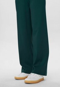 Pantalons verts à large jambe en tissu doux, associés à des baskets blanches avec une semelle en caoutchouc brun clair. Texture lisse et coupe décontractée.