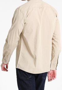 Camicia beige a maniche lunghe con colletto button-down, cucitura della spalla posteriore e orlo arrotondato. Presenta polsini con bottoni e un tessuto leggero e testurizzato.