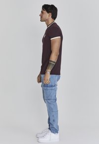 Bordeaux t-shirt met contrasterende crèmekleurige kraag en mouwen, gecombineerd met lichtblauwe cargobroek en witte sneakers, zichtbaar vanaf de zijkant.