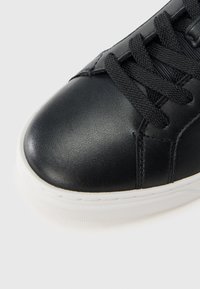 Mustast nahast toss, millel on mustad nöörid ja valge kummist tall, näidates suurt plaani varba ja paelte ala tavalise taustaga.