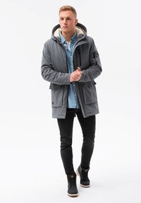 Grå vattentålig jacka med en vit fleecefoderhuva, dragkedja fram, två nedre fickor, bärs över en ljusblå skjorta och svarta jeans. Svarta stövlar.