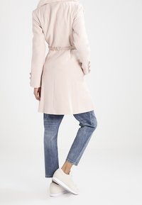 Cappotto rosa chiaro con colletto di pelliccia, vita fermata da una cintura, maniche lunghe e polsini bottonati, abbinato a jeans in denim blu e sneakers chiare.