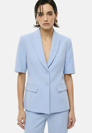 Donna con capelli corti e scuri, che indossa orecchini a cerchio dorati e una giacca da completo azzurro chiaro a maniche corte con pantaloni abbinati su uno sfondo uniforme.