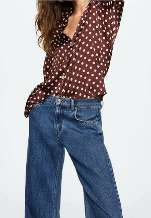 Kobieta w brązowej koszuli w kropki zapinanej na guziki, włożonej do wysokich, szerokich dżinsów w kolorze niebieskim, na białym tle.