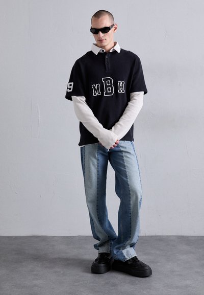 Schwarzes Polo mit kurzen Ärmeln, weißen Akzenten und Text, übereinander getragen mit einem langen weißen Basisteil, kombiniert mit hellblauen Jeans und schwarzen Sneakers.