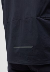 Giacca blu navy con una texture liscia, caratterizzata da una sottile striscia riflettente vicino all'orlo e un design pulito e minimale.