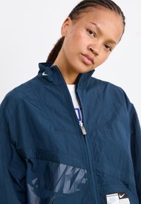 Jūras zils vējjakas ar augstu apkakli, pusčaklu, paneļu dizainu un spīguļojošu Nike logotipu. Izgatavota no teksturēta auduma un ar sānu kabatu.
