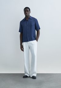 Mann kledd i marineblå kortermet skjorte med knapper, hvite vide bukser og svarte sko, stående mot en ensfarget lysegrå bakgrunn.