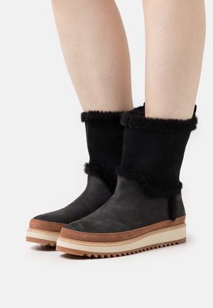 Bottes noires mi-mollet avec bordure en fourrure noire, détail en daim marron au-dessus des semelles plates crème avec semelles extérieures marron striées sur jambes nues.