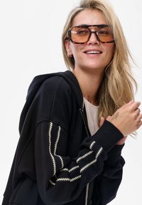 Czarna bluza z kapturem z białymi szczegółami w zygzakowate szycie, z zamkiem z przodu i sznurkiem do ściagania kaptura. Duże okulary przeciwsłoneczne w muszelkowej oprawie z brązowymi szkłami.