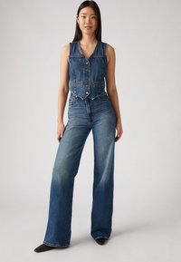 Denimvest og bukser med vide ben i medium blå vask. Vesten har knappelukking og flettet søm; buksene har høy midje og lett falming.