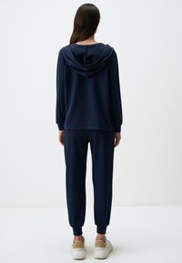 Completo felpa blu navy composto da felpa con cappuccio e pantaloni jogger realizzati in tessuto morbido; presenta un cappuccio con cordino, polsini a coste e orli alle caviglie. Scarpe da ginnastica visibili sotto.