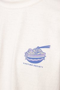 Biela bavlnená tričko s modrou grafikou misy rezancov, kreviet a paličiek na jedenie. Pod dizajnom je text "KAOTIKO SOCIETY".