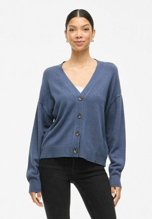 Femme aux cheveux foncés portant un cardigan bleu à boutons et un pantalon noir, se tenant devant un fond blanc uni.