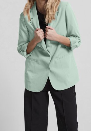 Femme aux cheveux blonds portant un blazer vert clair sur un haut noir et un pantalon noir, tenant les revers du blazer avec les deux mains.