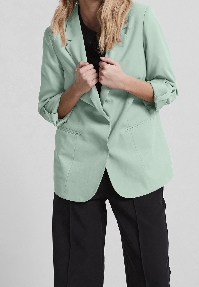 Femme aux cheveux blonds portant un blazer vert clair sur un haut noir et un pantalon noir, tenant les revers du blazer avec les deux mains.