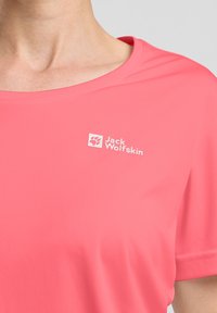 Camicetta rosa a maniche corte realizzata in tessuto leggero. Presenta un collo rotondo e un logo bianco sul petto. Texture liscia, design casual.