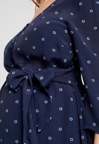 Mörkblå klänning med v-ringning, långa ärmar och bälte; har små vita blommönster och en mjuk, slät textur.