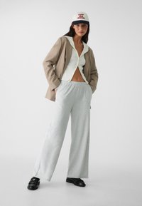 Blazer beige, haut en tricot crème à zip côtelé, pantalon de survêtement gris à large jambe, mocassins noirs et une casquette blanche avec du texte rouge. Tenue simple et décontractée.