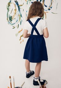 Dieťa s dlhými vlasmi, oblečené v bielej košeli a námorníckom modrom plášti, stojí a maľuje abstraktné farebné ťahy na bielu stenu.