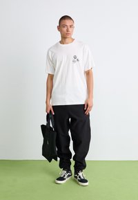Tricou alb din bumbac cu grafica mică neagră, purtat cu pantaloni negri și o geantă tote neagră. Modelul stă pe un podea verde.