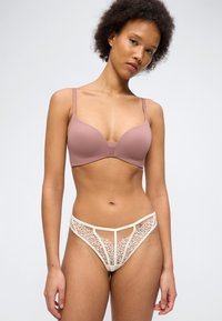 Γυναίκα με σγουρά μαλλιά που φοράει μωβ σουτιέν και μπεζ δαντελένιο σλιπ με ψηλή κοπή, στέκεται μπροστά σε απλό λευκό φόντο.