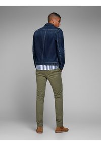 Chaqueta de mezclilla en azul oscuro con un diseño estructurado, llevada sobre una camisa a rayas. Pantalones verde olivo y zapatos color tostado completan el conjunto.