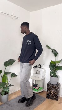 Sweatshirt preto com gráfico frontal, jeans cinza de perna larga e sapatos pretos. Ambiente interior com plantas verdes e uma máquina de costura.