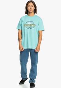 Turquoise t-shirt with "Quiksilver" logo in black and yellow, paired with blue jeans and black shoes. Short sleeves and relaxed fit.