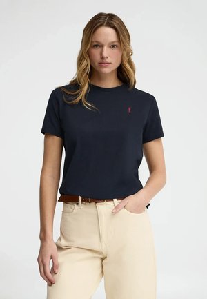 Frau mit welligem blondem Haar trägt ein marineblaues T-Shirt mit kurzen Ärmeln und beige Hose mit braunem Gürtel, sie steht vor einem schlichten hellen Hintergrund.