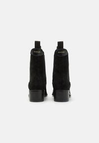 Botas de tobillo de ante negro con tacón redondeado, que presentan dos lengüetas y un detalle de logo dorado en el cuello. Textura suave, diseño elegante.