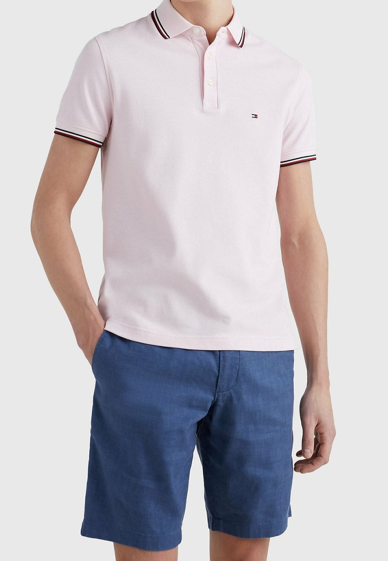Polo rose clair avec une patte de boutonnage à deux boutons et des accents rayés noir/rouge sur le col et les manches, assorti à un short bleu.