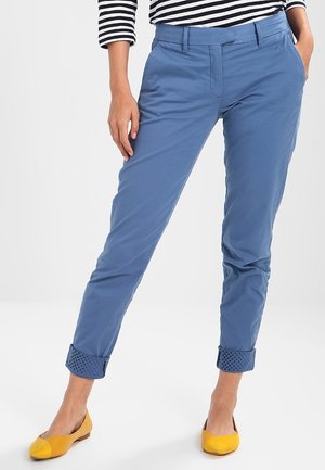 Vrouw met blauwe opgerolde broek, gele platte schoenen en een zwart-wit gestreept shirt met lange mouwen, staand met één hand in de zak.