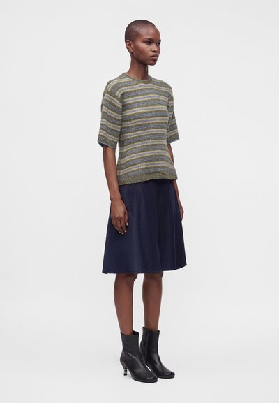 Femme portant un pull à manches courtes rayé, une jupe trapèze bleu marine et des bottines noires, debout devant un fond clair uni.