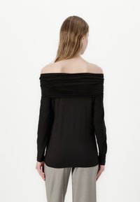 Black off-the-shoulder top with long sleeves, featuring gathered fabric around the neckline and a smooth texture. Styled with neutral trousers.