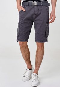 Grå cargoshorts med flera fickor, ett bälte och synliga söm detaljer, kombinerat med vita sneakers på en enkel bakgrund.