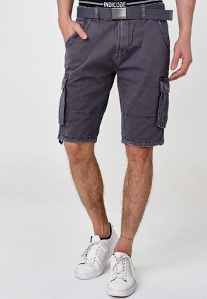 Shorts cargo gris avec plusieurs poches, une ceinture et des détails de couture visibles, portés avec des baskets blanches sur un fond uni.