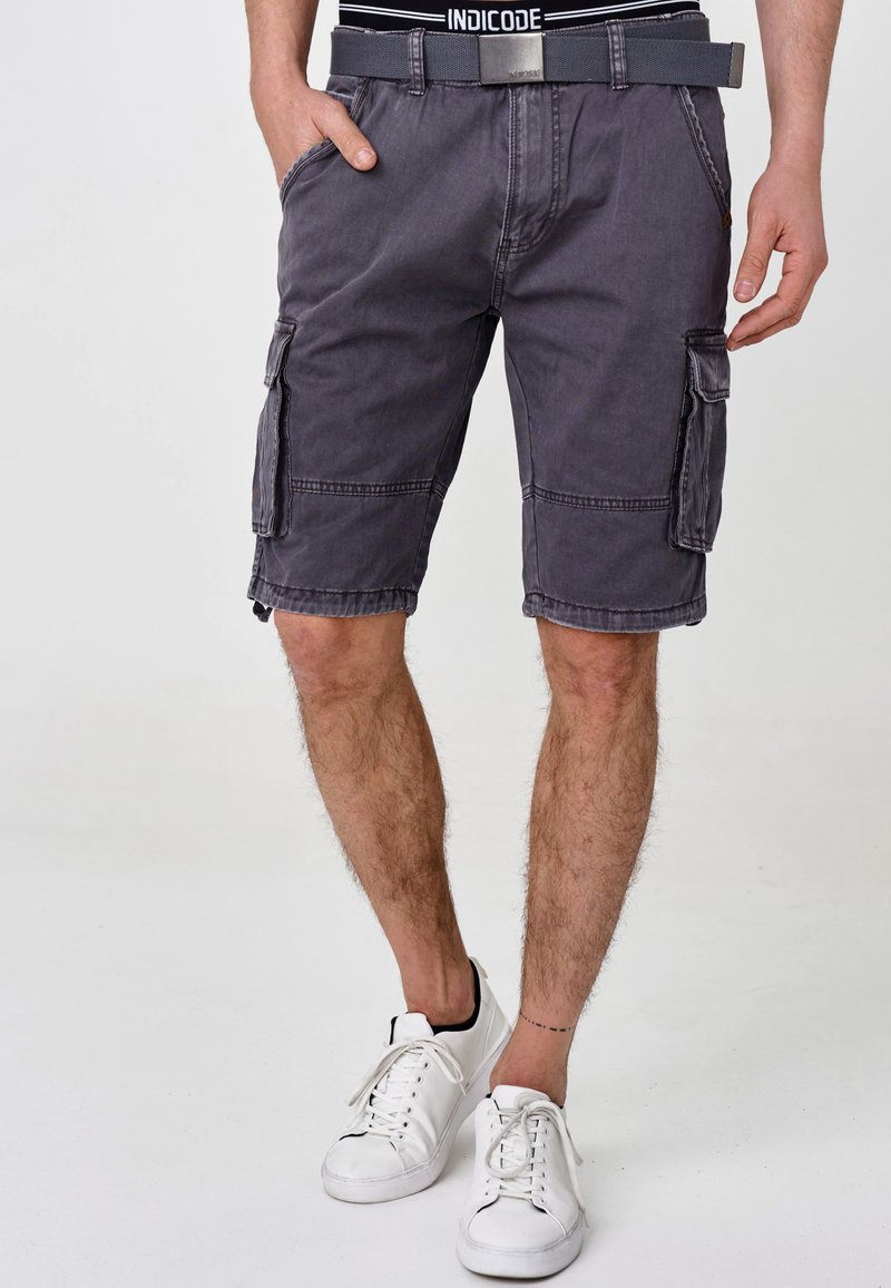 Grå cargo-shorts med flere lommer, belte og synlige sømdetaljer, kombinert med hvite joggesko på en enkel bakgrunn.