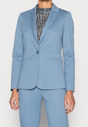 Femme portant un blazer bleu clair cintré avec un seul bouton et un pantalon assorti sur un top noir et gris à col montant et motif.