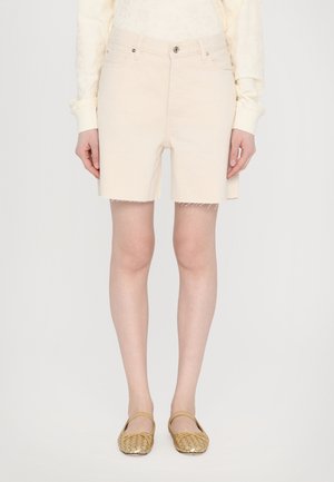 Personne portant un short en denim couleur crème avec des ourlets bruts, un haut léger à manches longues et des chaussures plates tissées dorées, debout devant un fond uni.