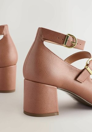 Różowe skórzane buty na słupku z paskiem wokół kostki i złotym zapięciem. Posiadają wycięty wzór oraz teksturowane wykończenie cholewki.