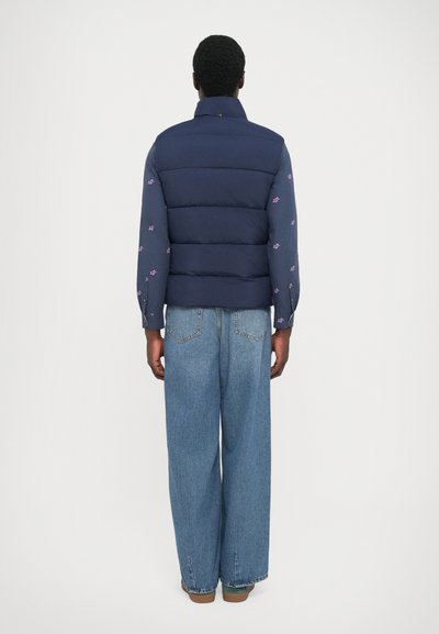 Navy puffer vest over a floral long-sleeve shirt, paired with baggy light blue jeans and brown shoes, viewed from the back.