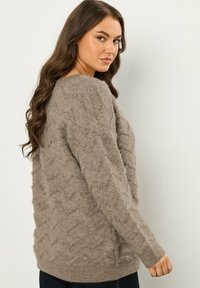 Puszysty sweter w kolorze taupe z teksturą w zygzak i ściągaczami przy rękawach, o luźnym kroju i długich rękawach, pokazany od tyłu.