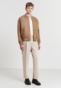 Giovane uomo con giacca bomber beige, camicia crema, pantaloni beige e mocassini marrone scuro in piedi contro uno sfondo bianco.